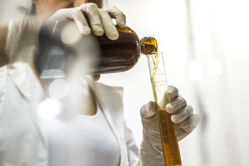 Mulher usando luvas descartáveis despeja um liquido amarelo de um compartimento de vidro para um tubo de ensaio.