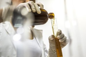Mulher usando luvas descartáveis despeja um liquido amarelo de um compartimento de vidro para um tubo de ensaio.