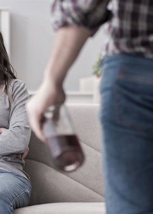 Homem segurando garrafa de bebida e mulher de fundo sentada no sofá
