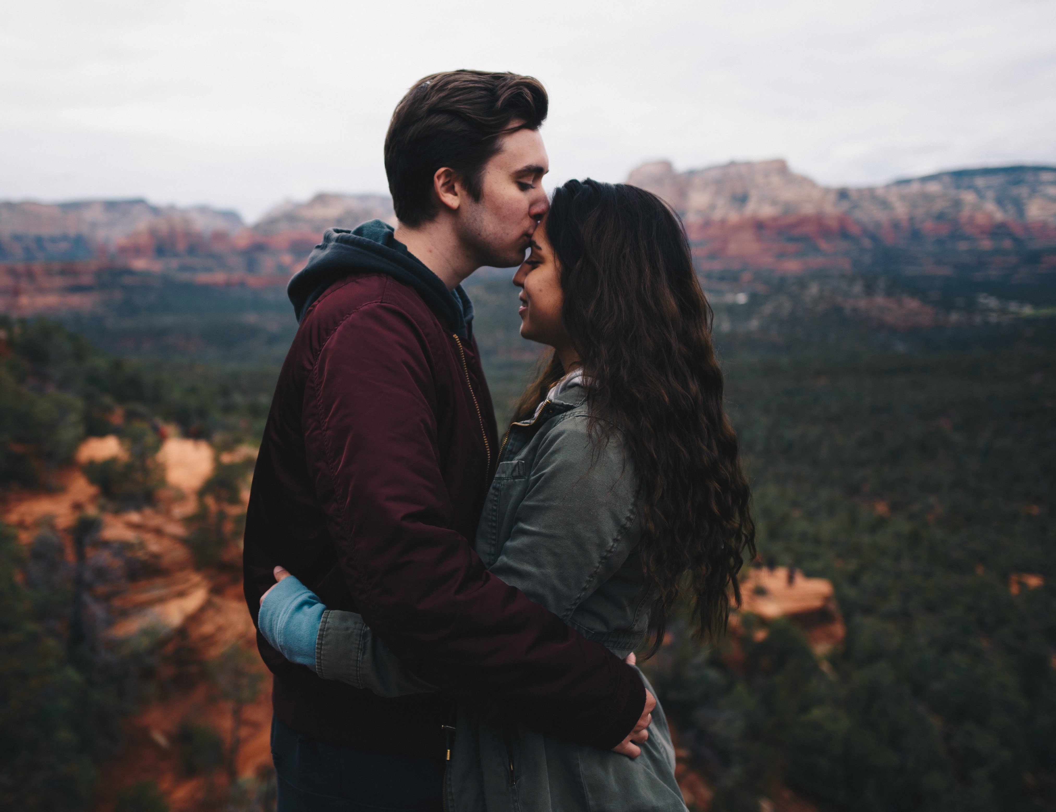 Homem beijando cabeça da mulher enquanto estão abraçados no topo de uma montanha