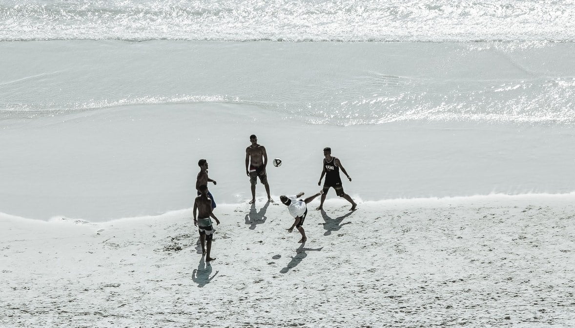 Grupo de cinco homens jogando futebol na praia, perto da água. Foto em preto e branco.