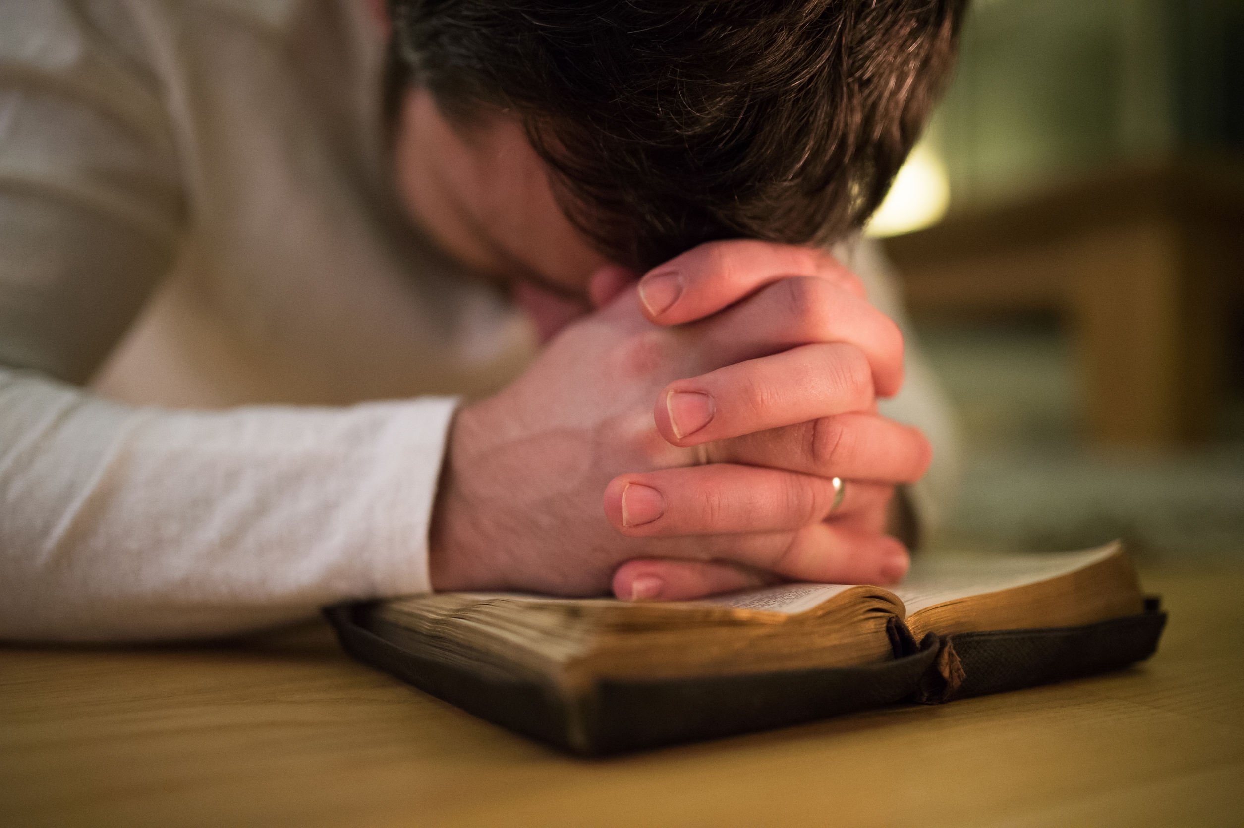 Homem jovem as mãos entrelaçadas apoiando em uma bíblia aberta, e sua cabeça baixa se apoia em suas mãos.