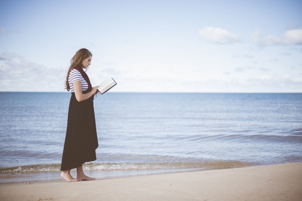 Mulher jovem com vestido andando na praia próxima ao mar, lendo uma bíblia.