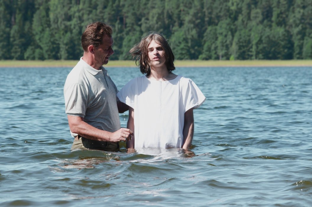 Menino vestindo branco, ao lado de homem adulto, ambos dentro de um rio. O menino está sendo batizado.