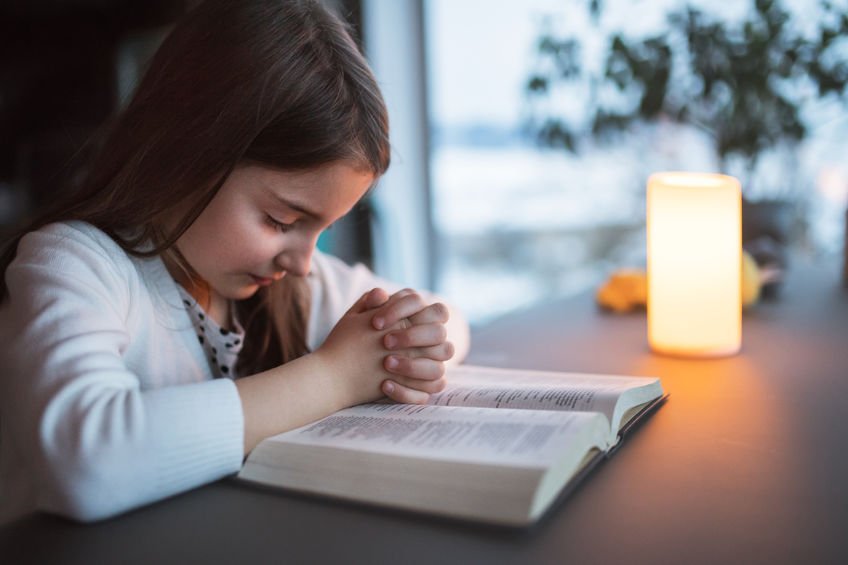 Menina com os olhos fechados as mãos entrelaçadas em cima de uma bíblia aberta.