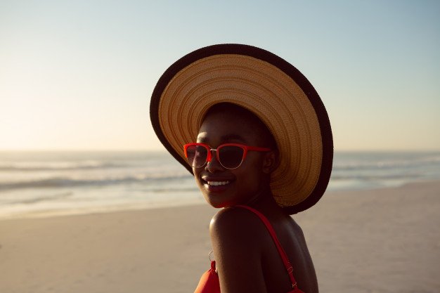 Mulher de chapéu e óculos sorrindo na praia.