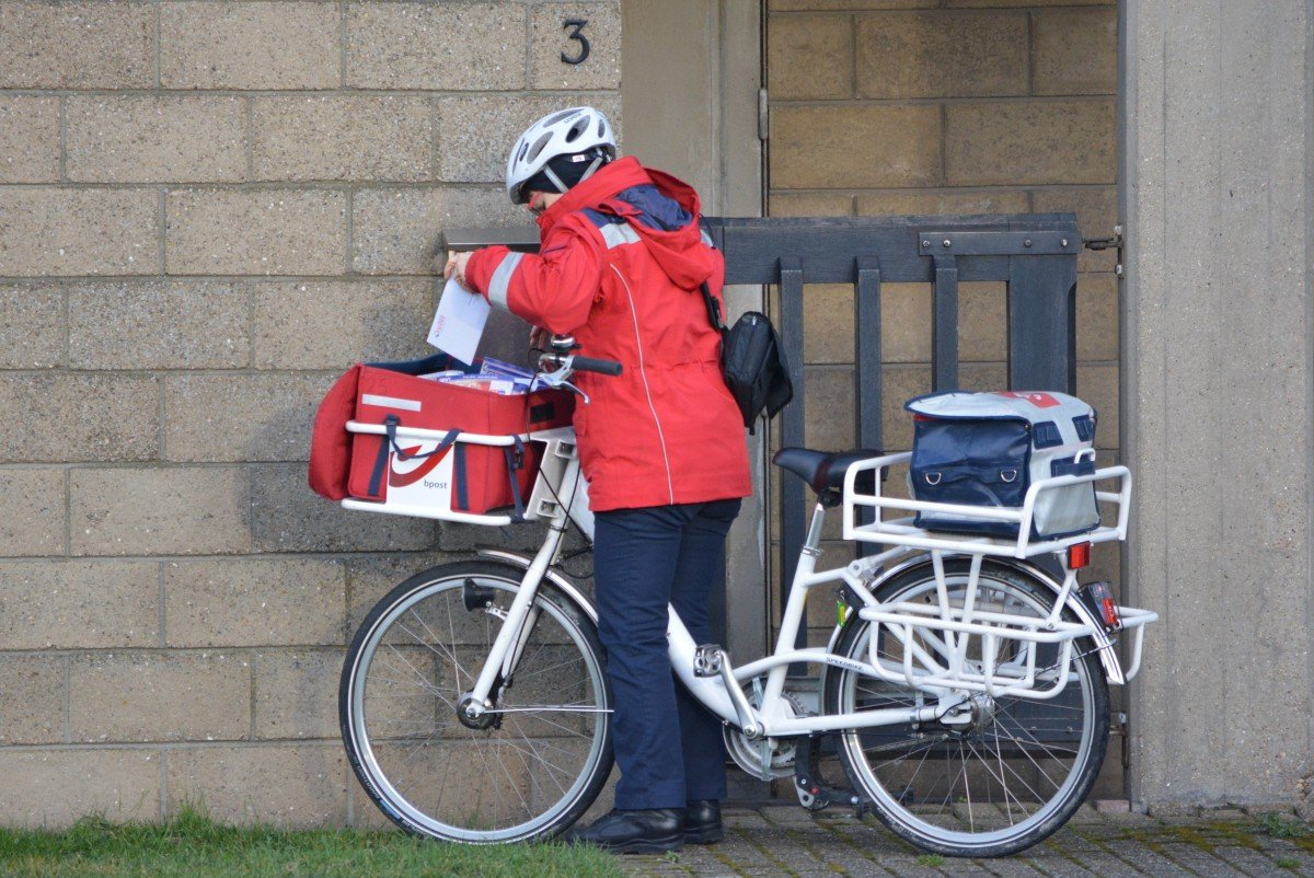 Carteiro com bicicleta e uniforme vermelho parado na porta de uma casa, e tirando um envelope de dentro de uma mochila vermelha.