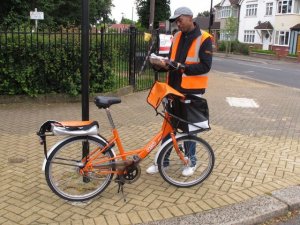 Carteiro com bicicleta e uniforme laranja olha uma pilha de cartas em suas mãos.