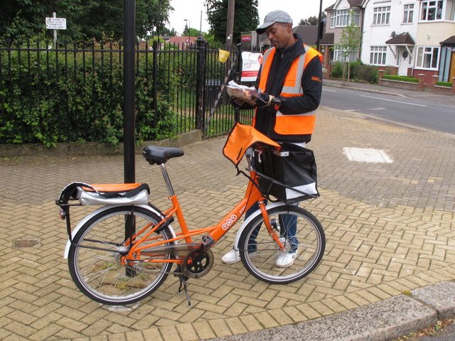 Carteiro com bicicleta e uniforme laranja olha uma pilha de cartas em suas mãos.