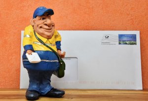 Boneco miniatura de carteiro sorrindo e entregando carta, com um envelope de carta em tamanho real ao fundo.