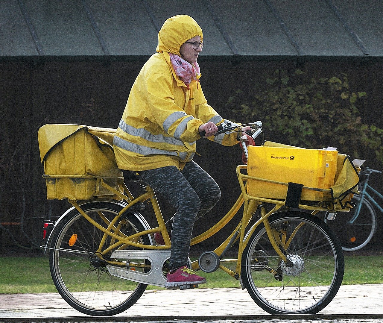 Carteira com jaqueta amarela em bicicleta pedalando. Na parte de trás da bicicleta, uma bolsa está presa. Na parte da frente da bicicleta, uma cesta contém diversos pacotes.
