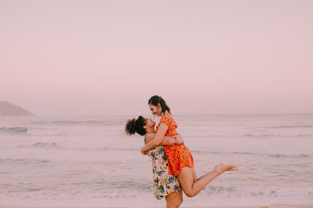 Mulher branca e mulher negra se abraçando numa praia.