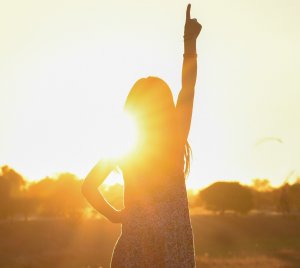 Por: Jude Beck Mulher de frente para o por do sol com o braço sinalizando o céu