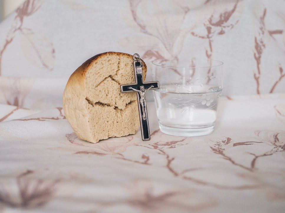 Pedaço de pão, copo transparente com água, e um pingente de Jesus crucificado na frente dos dois.