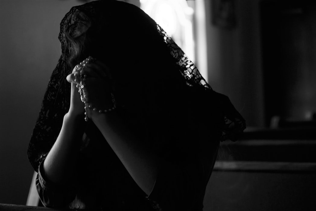 Mulher rezando com a cabeça coberta por um véu preto, segura um terço com suas mãos, que estão entrelaçadas e encostando em sua testa.