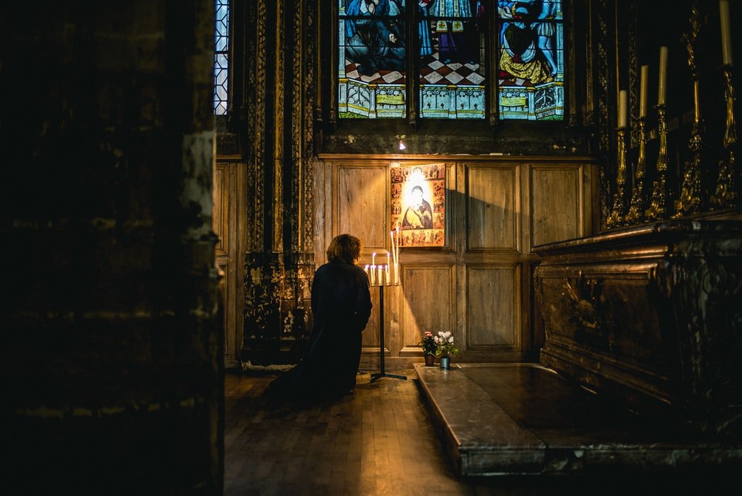 Mulher ajoelhada em igreja, de frente para uma imagem e velas acesas.