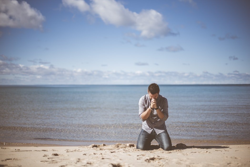 Homem ajoelhado em praia, na areia, rezando.
