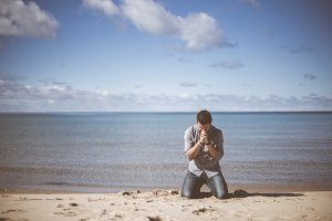 Unsplash - Ben White Homem ajoelhado em praia, na areia, rezando.