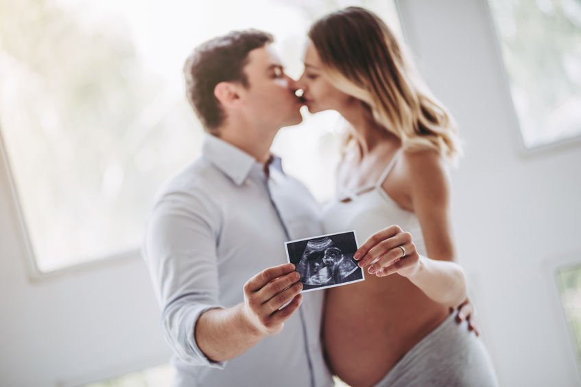 Homem e mulher gestante se beijando e segurando uma foto de ultrassom.