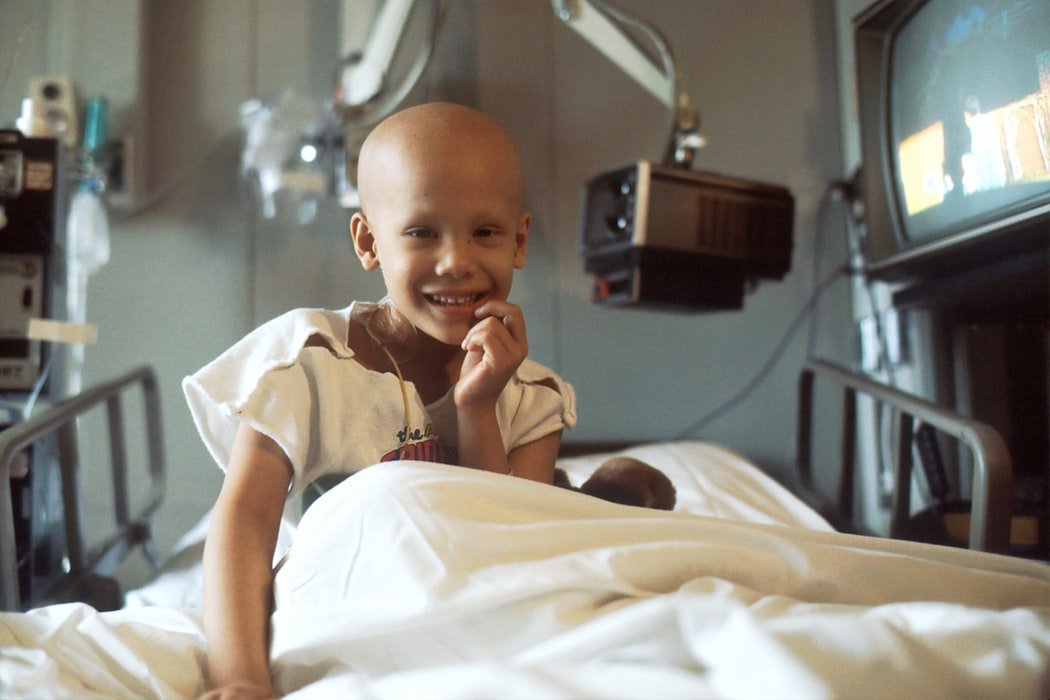 Criança com câncer sorrindo e sentada em uma maca de hospital.