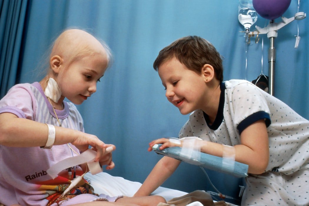 Criança com câncer e criança com o braço quebrado brincam em hospital.