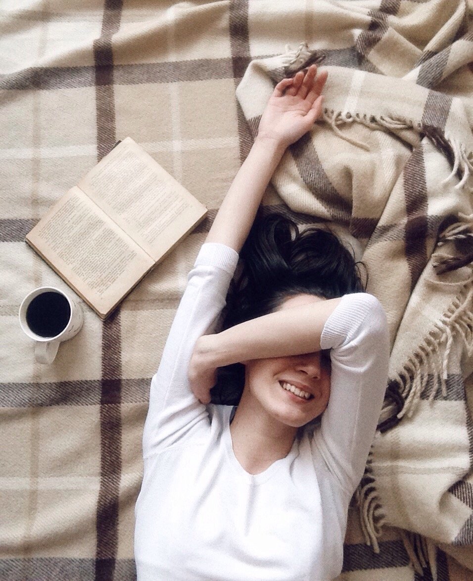 Mulher deitada na cama sorrindo com uma xícara de café e um livro ao seu lado