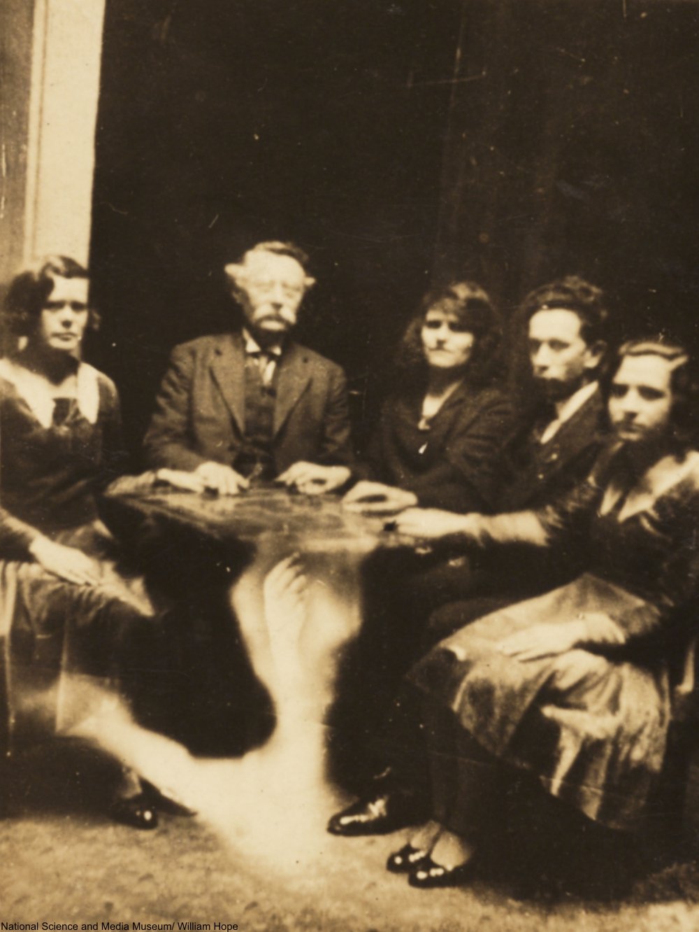 Foto de reunião espírita no meio do século XIX. Pessoas sentadas ao redor de uma mesa, com as mãos em cima da mesa.