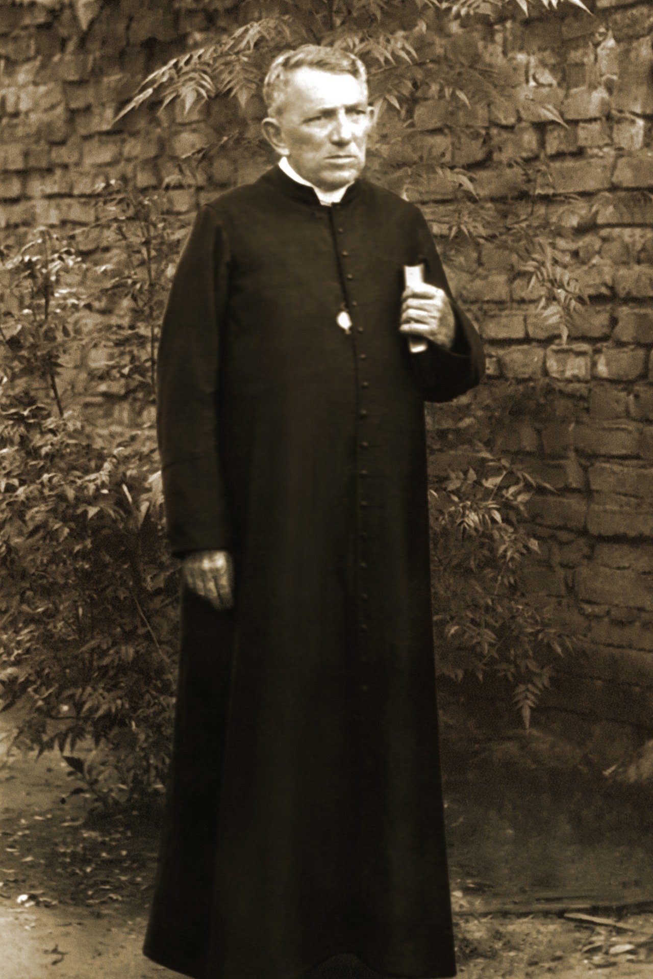 Padre Cícero de pé, em frente a um muro de tijolos, segurando uma bíblia embaixo do braço esquerdo.
