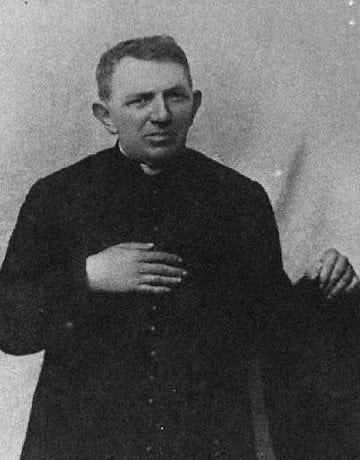 Padre Cícero de pé, com a mão direita apoiada no peito, e sua mão esquerda apoiada em uma cadeira.