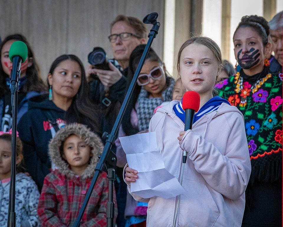 Greta Thunberg falando em microfone durante um protesto, com diversos manifestantes atrás dela.