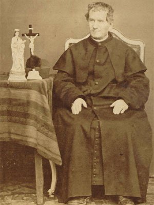 Colégio Salesiano Santa Teresinha São João Bosco sentado ao lado de uma mesa com duas estátuas, uma de Jesus na cruz, e a outra de Maria com Jesus no colo.