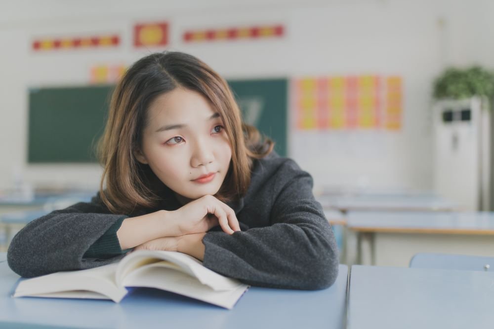 Garota em sala de aula apoiada em cima de um livro com expressão de pensativa.