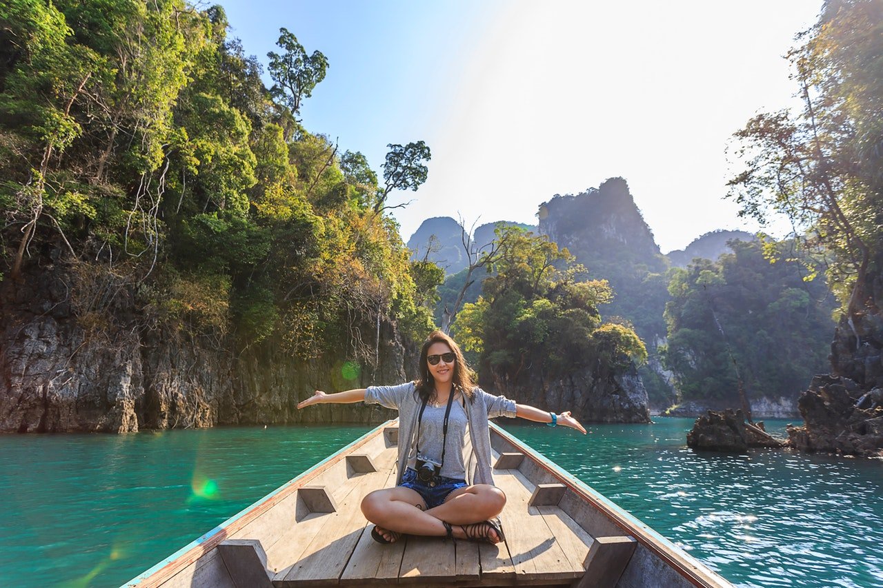 Mulher dentro de um barco, posando para foto com os braços abertos. Ela está em um local com água azul esverdeada e muitas ilhas montanhosas com vegetação.