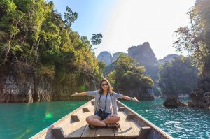 Pexels - Te lensFix Mulher dentro de um barco, posando para foto com os braços abertos. Ela está em um local com água azul esverdeada e muitas ilhas montanhosas com vegetação.