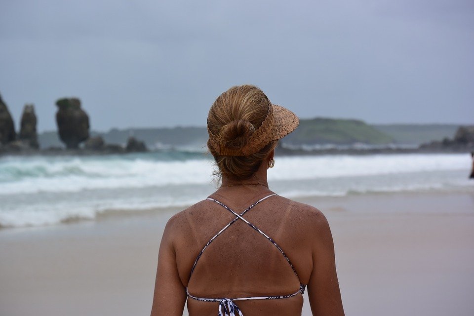 Mulher usando biquini, caminhando numa praia em Fernando de Noronha. Ela está de costas para a câmera e é possível ver o mar e ilhas rochosas na frente dela.