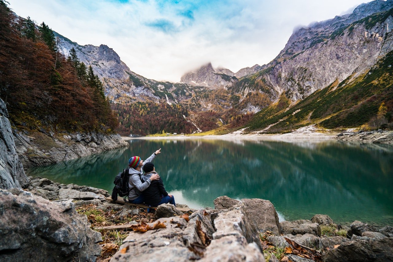 Duas pessoas com roupas de inverno sentadas em pedras na beira de um lago, rodeado por montanhas.