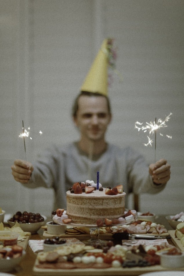 Homem aniversariante segura velas acesas atrás da mesa do bolo.