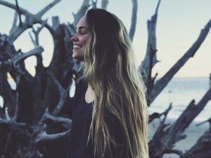 Rachel Gagnon Reshot Mulher sorrindo na praia ao lado de uma árvore seca