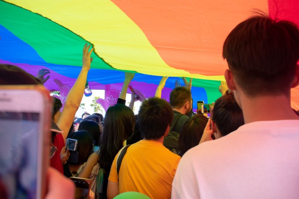 Multidão embaixo de uma grande bandeira com as cores do arco-íris.