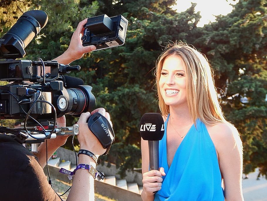 Mulher segurando microfone e sorrindo para uma filmadora profissional que está em sua frente.