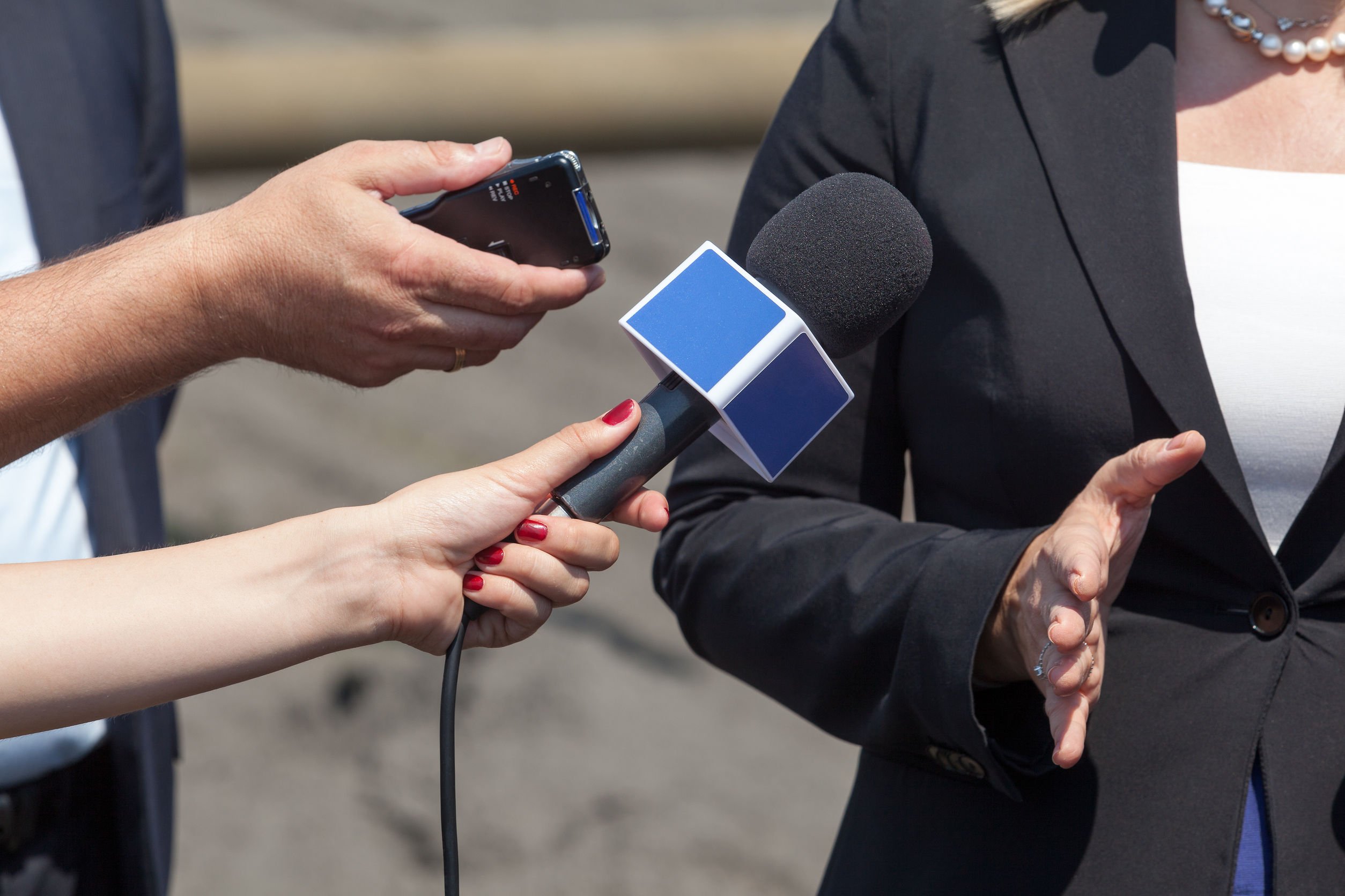 Duas mãos de pessoas diferentes, uma segurando um gravador portátil e a outra segurando um microfone, apontam seus equipamentos para uma mulher que está falando.