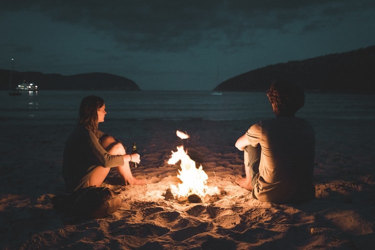 Casal sentado em areia de praia ao lado de fogueira à noite