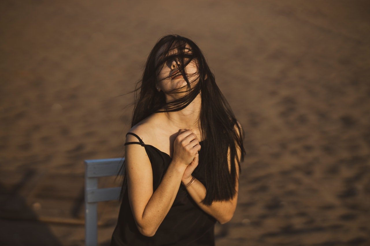 Mulher sentada numa praia, com o cabelo no rosto devido ao vento, e as mãos unidas em frente de seu peito.