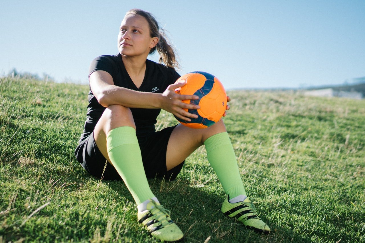 Mulher sentada em gramado com os joelhos flexionados. Ela olha para o lado enquanto segura uma bola de futebol em frente ao seu corpo.