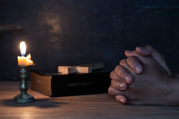 Mãos juntas rezando, próxima a uma vela e uma bíblia.