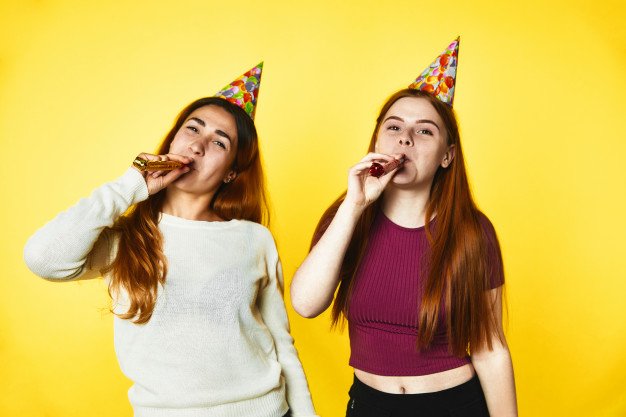Duas garotas com chapéu de aniversário assoprando língua-de-sogra.