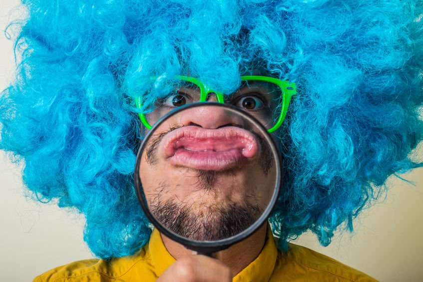Homem com peruca azul e óculos neon segurando lupa em frente a boca
