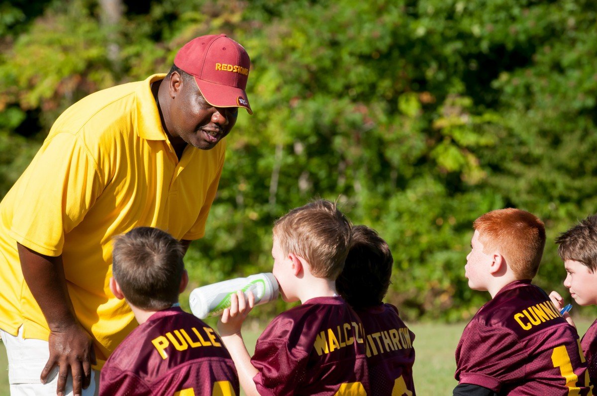Treinador usando camiseta amarela e boné de seu time, falando com seus jogadores, pequenos meninos com uniforme vermelho do time.