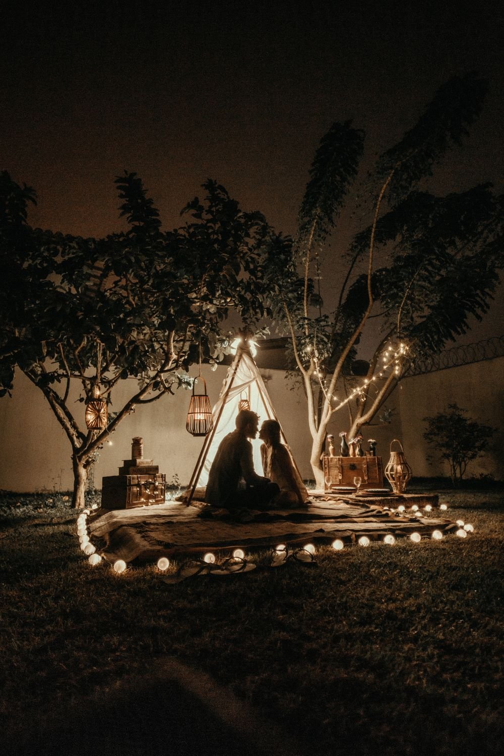 Casal acampado ao lado de árvores, com velas e iluminações em volta.