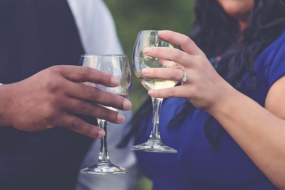 Casal brindando com taças cheias de vinho branco. A mulher usa um anel de noivado no dedo anelar esquerdo.